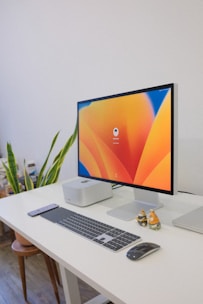 A clean and organized workspace featuring a modern desktop computer. The computer monitor displays a colorful abstract design as the screensaver. The setup includes a wireless keyboard, a wireless mouse, and a smartphone placed on a white desk. To the left of the monitor, there is a small computer unit and a plant, adding a touch of greenery to the environment. To the right of the monitor, there are two small decorative figurines.