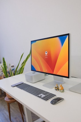 A clean and organized workspace featuring a modern desktop computer. The computer monitor displays a colorful abstract design as the screensaver. The setup includes a wireless keyboard, a wireless mouse, and a smartphone placed on a white desk. To the left of the monitor, there is a small computer unit and a plant, adding a touch of greenery to the environment. To the right of the monitor, there are two small decorative figurines.