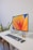A clean and organized workspace featuring a modern desktop computer. The computer monitor displays a colorful abstract design as the screensaver. The setup includes a wireless keyboard, a wireless mouse, and a smartphone placed on a white desk. To the left of the monitor, there is a small computer unit and a plant, adding a touch of greenery to the environment. To the right of the monitor, there are two small decorative figurines.