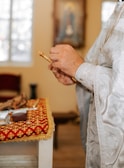 A person dressed in ornate, decorative white and silver robes holding a small golden object. The background includes a table covered with a richly decorated red and gold cloth, with various items including books and a smaller red object. The setting appears religious or ceremonial.