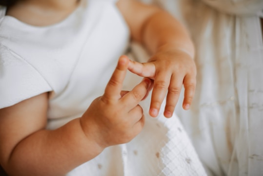 A child wearing a white dress is delicately touching the fingers on one hand with the other. The focus is on the hands, showcasing soft, youthful skin.