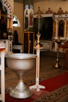 A church interior with traditional, ornate decorations featuring religious icons and paintings. In the foreground, there is a large silver baptismal font and a tall candle holder adorned with gold accents. The floor is covered with a detailed carpet in warm colors.