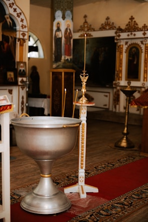 A church interior with traditional, ornate decorations featuring religious icons and paintings. In the foreground, there is a large silver baptismal font and a tall candle holder adorned with gold accents. The floor is covered with a detailed carpet in warm colors.