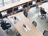 A modern office space with long wooden tables, several black ergonomic chairs, and a row of high stools by a window. A laptop, a cup of coffee, and some stationery are neatly arranged on one of the tables.