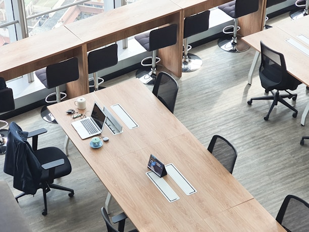 A modern office space with long wooden tables, several black ergonomic chairs, and a row of high stools by a window. A laptop, a cup of coffee, and some stationery are neatly arranged on one of the tables.