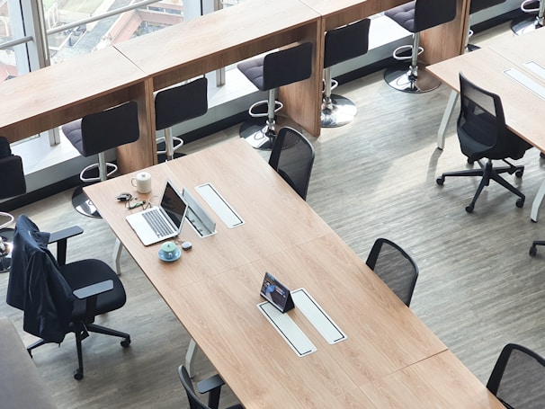 A modern office space with long wooden tables, several black ergonomic chairs, and a row of high stools by a window. A laptop, a cup of coffee, and some stationery are neatly arranged on one of the tables.