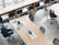 A modern office space with long wooden tables, several black ergonomic chairs, and a row of high stools by a window. A laptop, a cup of coffee, and some stationery are neatly arranged on one of the tables.