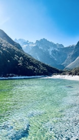 Crystal clear water of a lake with a vibrant green hue stretches into the distance, bordered by a lush, forested mountain range under a clear blue sky. Snow-capped peaks are visible in the background, and a few people can be seen on the shore, adding a sense of scale and activity.