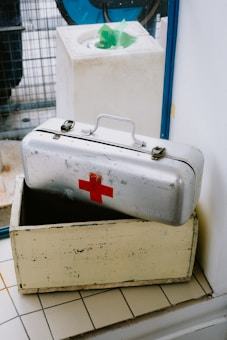 A metallic first aid kit marked with a red cross is positioned slightly open on top of a wooden box. It rests on a tiled floor next to a wall, with a background featuring a blue-framed glass panel and a white object with a green plastic lining.