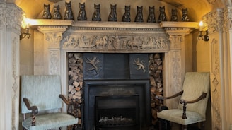 The image depicts a decorative stone fireplace adorned with intricate carvings, including several animal motifs and mythical creatures. A row of black cat figurines lines the mantel. Two ornate chairs with fabric upholstery are positioned on either side of the fireplace. Logs are stacked within the fireplace, and the setting is lit by wall-mounted lamps, contributing to an elaborate and historical ambiance.