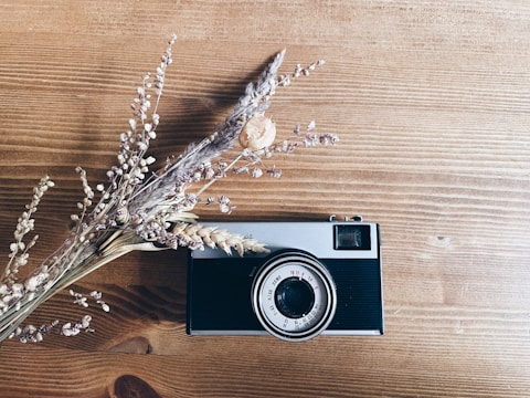 A vintage camera lies on a wooden surface next to a bundle of dried flowers, creating an aesthetically pleasing arrangement.