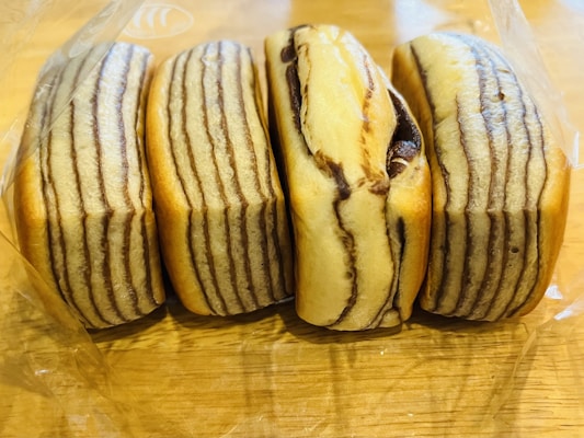Four loaf-like pastries are arranged side by side in a clear plastic bag on a wooden surface. They have a layered appearance with alternating light and dark brown stripes, giving a marbled effect.