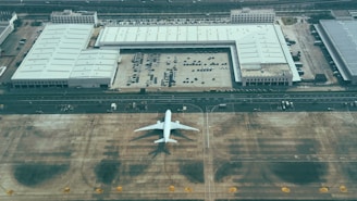 An aerial view displays a large aircraft parked on a tarmac next to expansive industrial buildings with multiple parked vehicles. The setting is a spacious area with marked runways and driving lanes, possibly indicating a maintenance or storage facility.