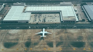 An aerial view displays a large aircraft parked on a tarmac next to expansive industrial buildings with multiple parked vehicles. The setting is a spacious area with marked runways and driving lanes, possibly indicating a maintenance or storage facility.
