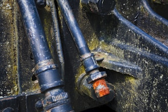 Dark metal pipes and bolts with evident rust and wear, mounted on an industrial or mechanical structure. The metal surfaces display signs of aging and corrosion, showing patches of yellowish-green mold or oxidation.