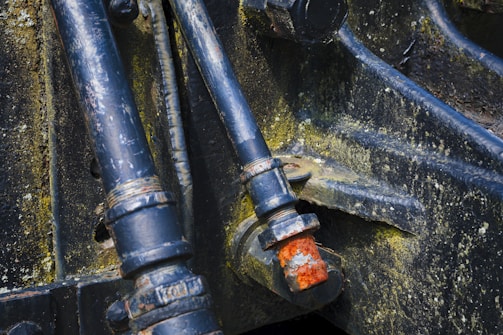 Dark metal pipes and bolts with evident rust and wear, mounted on an industrial or mechanical structure. The metal surfaces display signs of aging and corrosion, showing patches of yellowish-green mold or oxidation.