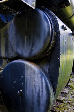 Close-up view of a large metal boiler or tank with a weathered surface. The cylindrical body shows signs of rust and wear, while bolts and rivets are visible along the edges.