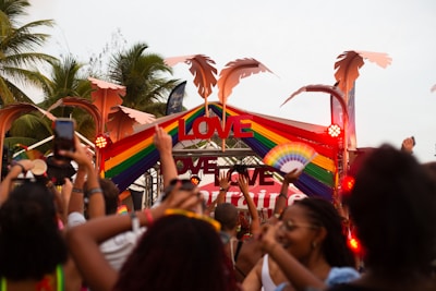 A crowd of people is enjoying an outdoor event, holding up their hands and electronic devices. The event has vibrant rainbow-themed decorations with the word 'LOVE' prominently displayed above the stage. Palm trees are visible in the background, suggesting a tropical location.