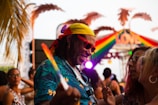 A vibrant outdoor scene with people enjoying a lively gathering. Prominent in the foreground is a man wearing a yellow bandana, red sunglasses, and a colorful tropical shirt, interacting joyfully with others. A rainbow-colored sign with 'LOVE' is visible in the background along with palm tree decorations, suggesting a festive and inclusive atmosphere.