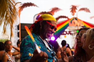A vibrant outdoor scene with people enjoying a lively gathering. Prominent in the foreground is a man wearing a yellow bandana, red sunglasses, and a colorful tropical shirt, interacting joyfully with others. A rainbow-colored sign with 'LOVE' is visible in the background along with palm tree decorations, suggesting a festive and inclusive atmosphere.