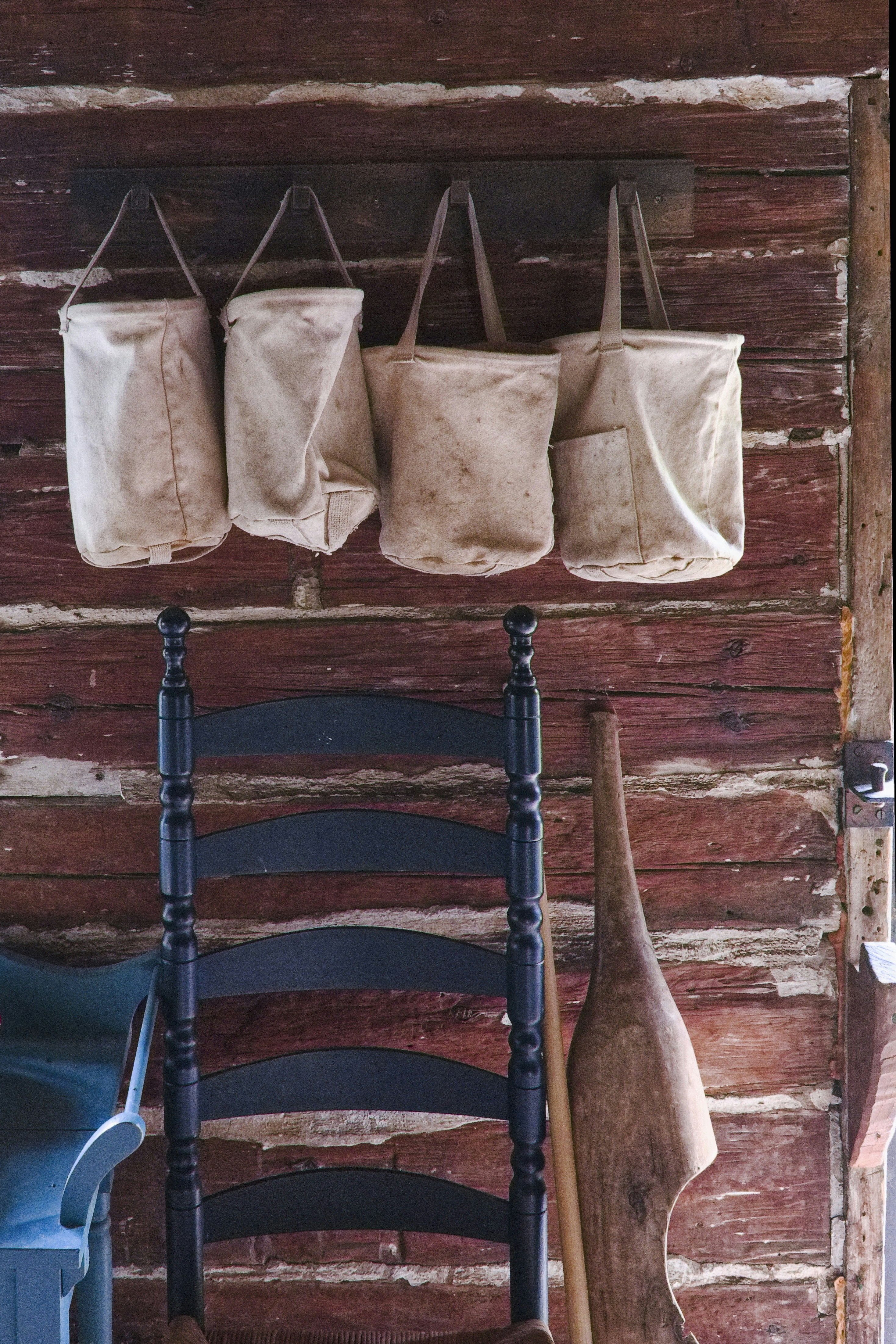 A rustic wooden wall features four canvas bags hanging on hooks. In front of the wall, part of a wooden chair is visible along with a carved wooden object leaning against the wall.