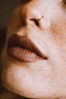 A close-up of a person's nose with a small hoop nose ring and full, matte lips. The skin is covered in freckles.