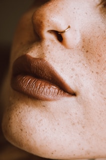A close-up of a person's nose with a small hoop nose ring and full, matte lips. The skin is covered in freckles.