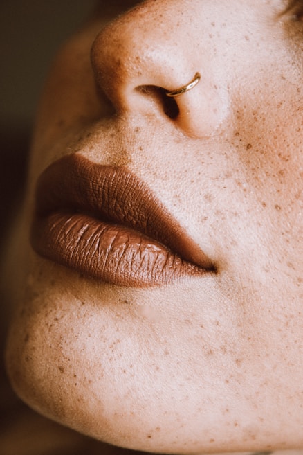 A close-up of a person's nose with a small hoop nose ring and full, matte lips. The skin is covered in freckles.