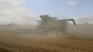 A large green combine harvester is operating in a dusty field of wheat. The machine is partially obscured by a cloud of dust as it moves through the harvested rows. The sky is mostly overcast with scattered clouds.