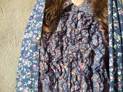 A close-up of a person wearing a blue ruffled shirt with a paisley pattern. The outer garment draped over the shoulders has a floral print with red, white, and green colors on a blue background. The hair is long and brown, draping over the shoulders.
