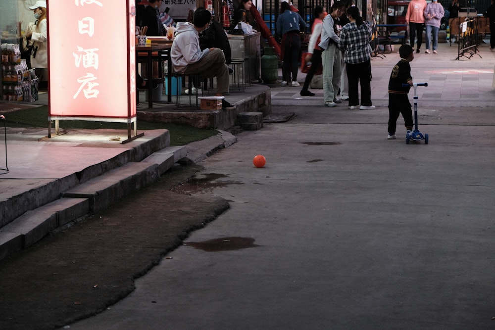 a young boy standing on a sidewalk next to an orange ball