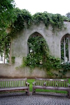 A serene and overgrown ruin with arched windows covered in lush green ivy. Two wooden benches are positioned on a cobblestone path, adding a peaceful atmosphere to the scene.