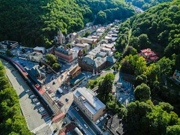 An aerial view of a small town nestled in a lush, hilly landscape. The town features a variety of buildings, including a church with a tall steeple, residential and commercial structures, and a visible train station. Cars are parked along streets and parking areas, and the image is surrounded by dense green foliage.