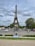 A young girl in a blue dress is sitting on a grassy area, facing the Eiffel Tower. The tower stands tall under a cloudy sky, and there are several people walking around and cars on the street nearby. Trees and lampposts are also visible, adding to the scenic urban setting.