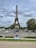 A young girl in a blue dress is sitting on a grassy area, facing the Eiffel Tower. The tower stands tall under a cloudy sky, and there are several people walking around and cars on the street nearby. Trees and lampposts are also visible, adding to the scenic urban setting.