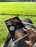 A hand holding a book titled 'Love in the Time of Cholera' by Gabriel García Márquez, while sitting on the grass in a bright, sunlit park. The cover features a portrait and a promotional sticker. The background shows a lush green field.