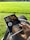 A hand holding a book titled 'Love in the Time of Cholera' by Gabriel García Márquez, while sitting on the grass in a bright, sunlit park. The cover features a portrait and a promotional sticker. The background shows a lush green field.