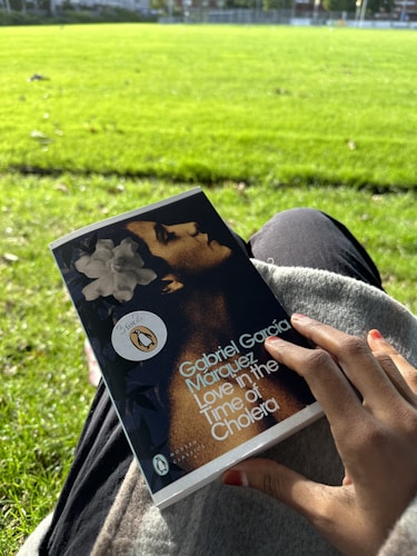 A hand holding a book titled 'Love in the Time of Cholera' by Gabriel García Márquez, while sitting on the grass in a bright, sunlit park. The cover features a portrait and a promotional sticker. The background shows a lush green field.