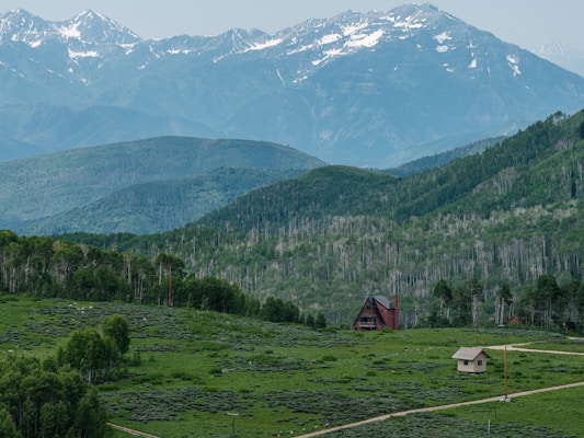 A scenic landscape featuring lush green hills with a dense coverage of trees and vegetation. In the background, there are majestic snow-capped mountains under a soft blue sky. A quaint A-frame house and a smaller structure are situated near the center of the image, adding a touch of human presence to the natural surroundings.