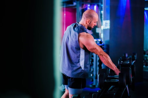 A man in a gym setting, wearing a sleeveless top and shorts, appears to be selecting a dumbbell from a rack. The environment is illuminated with soft, colorful lighting, creating a dynamic workout atmosphere.