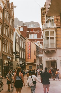 A bustling city street with several pedestrians walking in different directions. The street is lined with tall buildings featuring a mix of architectural styles. Visible storefronts include a casino with a vertical sign, a Bershka store, and other retail shops. The sky is overcast, adding a muted tone to the scene.