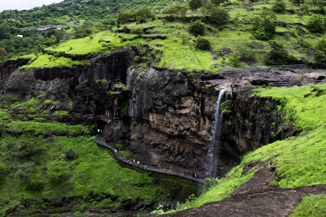 A lush green landscape features a cascading waterfall flowing down a rocky cliff. A narrow path with people walking along it meanders through the terrain. The area is surrounded by dense foliage and rolling hills.