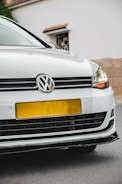 A close-up view of the front of a white Volkswagen car, featuring the emblem prominently displayed on the grille. The headlights are modern and sleek, and the car is parked on a street with a beige wall and a white building in the background.