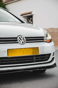 A close-up view of the front of a white Volkswagen car, featuring the emblem prominently displayed on the grille. The headlights are modern and sleek, and the car is parked on a street with a beige wall and a white building in the background.
