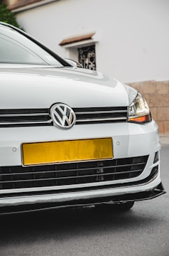A close-up view of the front of a white Volkswagen car, featuring the emblem prominently displayed on the grille. The headlights are modern and sleek, and the car is parked on a street with a beige wall and a white building in the background.