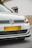 A close-up view of the front of a white Volkswagen car, featuring the emblem prominently displayed on the grille. The headlights are modern and sleek, and the car is parked on a street with a beige wall and a white building in the background.