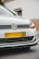 A close-up view of the front of a white Volkswagen car, featuring the emblem prominently displayed on the grille. The headlights are modern and sleek, and the car is parked on a street with a beige wall and a white building in the background.