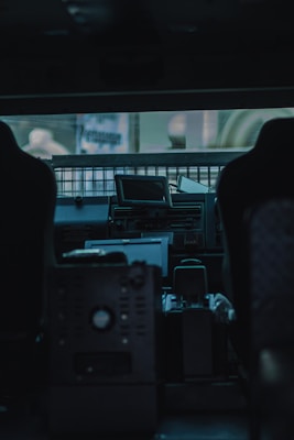The interior of a vehicle with various control panels and seats. The view includes a central dashboard area with several electronic devices and screens. Light filters through a wire mesh window, casting a muted glow over the scene.