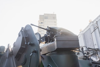A close-up view of military equipment, featuring the turret and gun barrel of an armored vehicle with a building visible in the background.