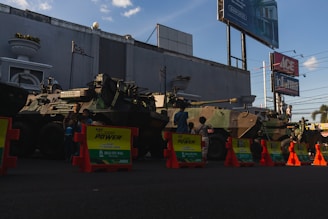 Two military armored vehicles are parked in a row on a street, partially obscured by orange and yellow traffic barriers in front. Several children stand around and climb on the vehicles. The background features a large building with minimalistic decoration, billboards, and power lines. The sky is clear with a few clouds.